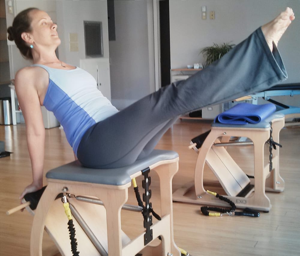 A white woman with her hair in a bun wearing a purple and lavender sports tank and grey flair yoga pants preforming pilates teaser on the chair.